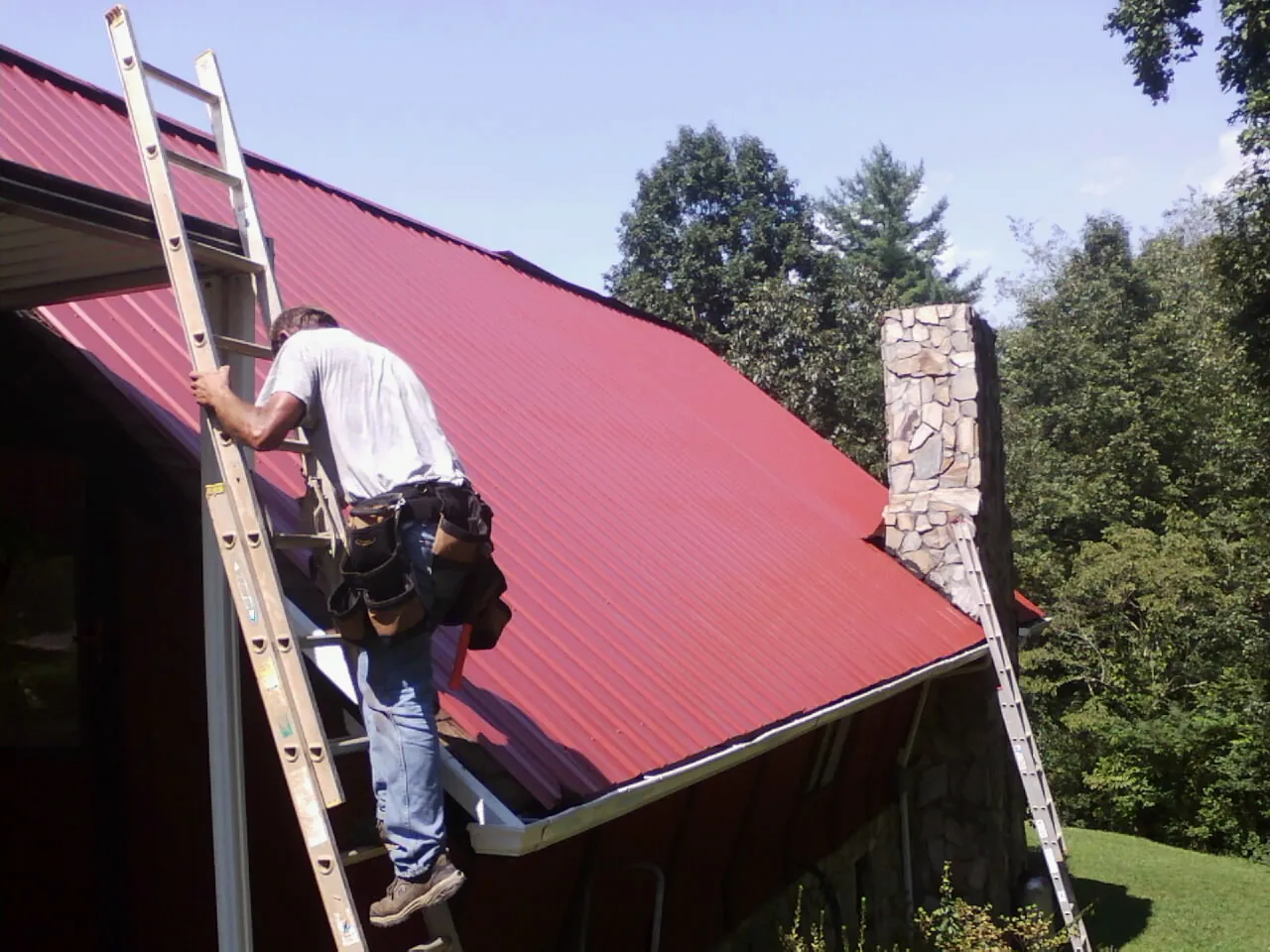 Roofer on a ladder beside a red metal roof and stone chimney — Soffit & Fascia Repair by Roofing Brothers in Mount Morris